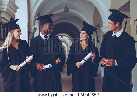 Happy to be graduated. Four college graduates in graduation gowns walking along university corridor and talking