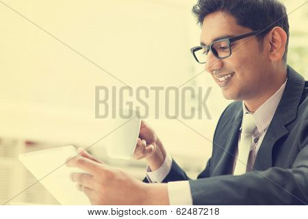 Young Asian Indian businessman using a tablet computer during office break at cafe, relaxing with a cup of coffee. India male business man,  in vintage retro style.