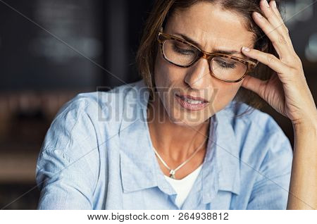 Portrait of stressed mature woman with hand on head looking down. Worried woman wearing spectacles. Tired lady having headache sitting indoors.