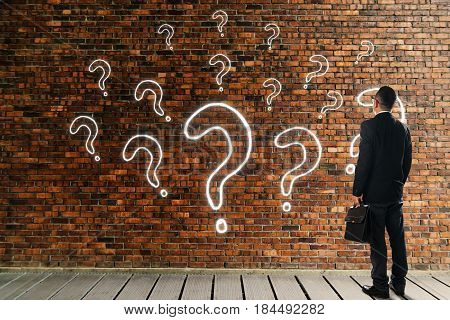 Businessman looking and thinking in front of a brick wall with the question marks icon . Business question concept .