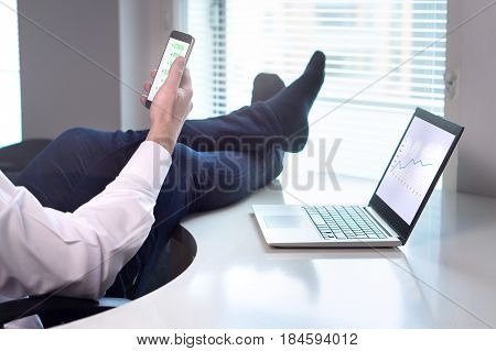 Successful business going well. Businessman laying in office with feet on table while income is rising and getting easy money. Stock market going up. Entrepreneur with smartphone and laptop in office.