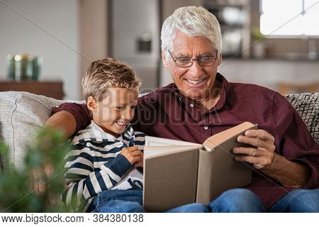 Grandfather and grandson reading a book and smiling. Happy little boy with old grandpa reading story book at home. Senior man tells a fairy tale story to his grandchild while sitting on couch.