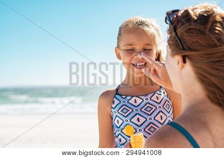 Young mother applying protective sunscreen on daughter nose at beach. Woman hand putting sun lotion on child face. Cute little girl with sunblock at seaside with copy space.