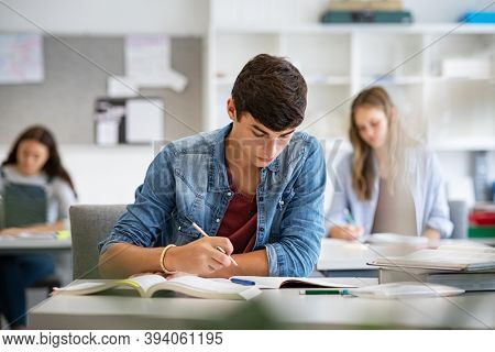 Focused young man taking notes from books for his study. College student sitting at desk and studying in high school library. Guy studying in classroom and completing project.