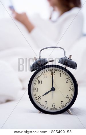 alarm clock standing on bedside table going to ring early morning to wake up woman in bed sleeping in background - early awakening -not getting enough sleep - getting work time concept