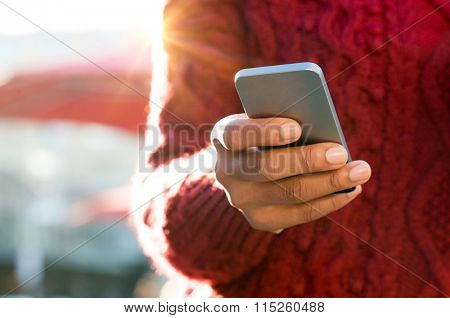 Close up of a young woman's hand typing text message on her smartphone. African young woman is typing on touch screen mobile phone. Close up of female hand texting a message phone outdoor. 
