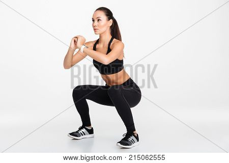 Portrait of a confident asian fitness woman doing squats isolated over white background