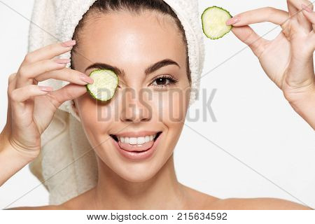 Close up beauty portrait of a smiling beautiful half naked woman with a towel wrapped around her face holding cucumber slices at her face and looking at camera isolated over white background