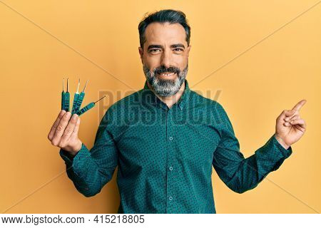 Middle age man with beard and grey hair holding picklock to unlock security door smiling happy pointing with hand and finger to the side 