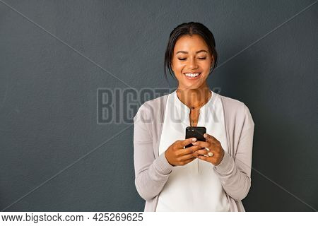 Mid adult african american woman texting message on smart phone isolated on grey background. Smiling indian woman using mobile phone. Happy multiethnic lady messaging on the smartphone with new app.