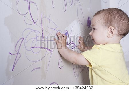 Baby boy drawing with wax crayon on plasterboard wall. He has the crayon on the mouth thinking what to do