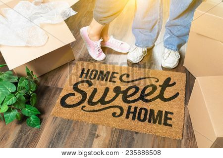 Man and Woman Standing Near Home Sweet Home Welcome Mat, Moving Boxes and Plant.