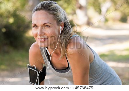 Portrait of athletic mature woman resting after jogging. Beautiful senior blonde woman running at the park on a sunny day. Female runner listening to music while jogging.