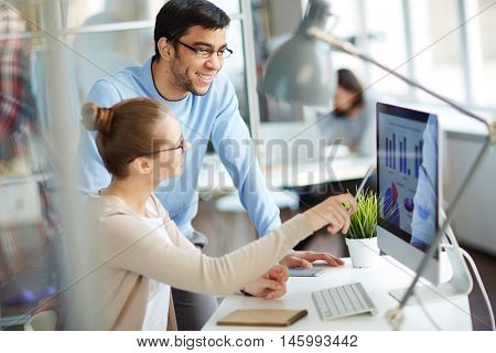 Two colleagues having business discussion in modern office, woman analyst showing successful financial and marketing statistics from screen to her male co-worker, sitting at desk with laptop computer