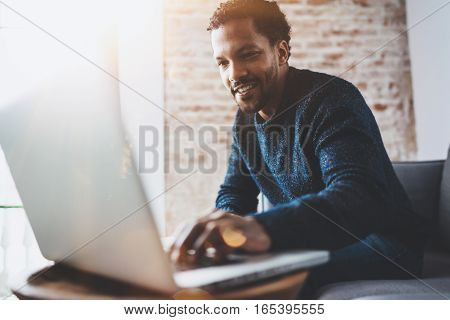 Cheerful African man using computer and smiling while sitting on the sofa. Concept of young business people working at home. Blurred background, flares