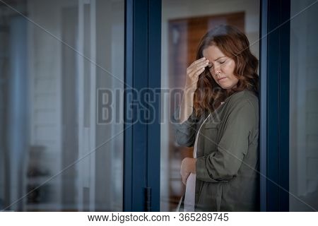 Stressed mature woman standing near window suffering from strong headache. Middle aged redhead woman with terrible migraine holding head with closed eyes. Mid lady feels pain in stomach, copy space.