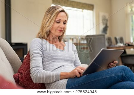 Beautiful mature woman sitting on couch and using digital tablet. Smiling lady browsing internet over tablet at home. Middle age woman holding computer at home.