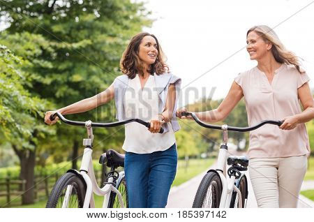 Two mature women in conversation while walking with bicycle at park. Happy beautiful senior women walking in the park with bicycles in a spring time. Friends holding bikes and talking to each other.
