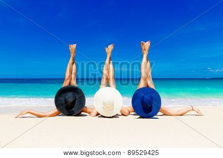 Three Young Women In Straw Hats Lying On A Tropical Beach, Stretching Up Slender Legs.