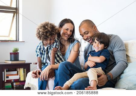 Happy multiethnic family sitting on sofa laughing together. Cheerful parents playing with their sons at home. Black father tickles his little boy while the mother and the brother smile.