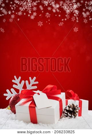 Close up of Christmas small box with red ribbon and empty greeting card. Xmas decoration with shiny ball and pine cone on snow. Christmas vertical red background.