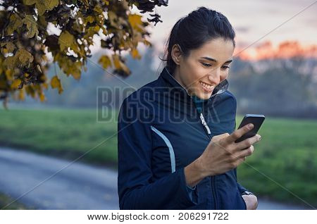 Young athlete looking at phone and smiling at park during sunset. Smiling young woman checking her smartphone while resting. Happy girl texting a phone message with a big smile on her face.