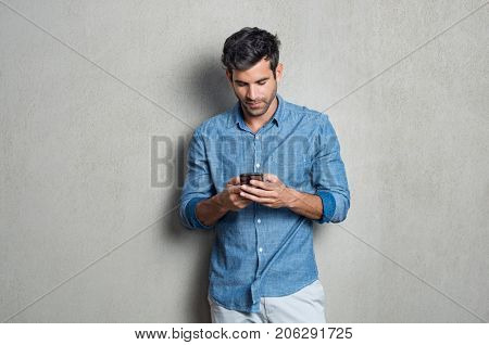 Young man writing phone message. Mid adult man typing on mobile while standing on grey background. Latin man leaning on gray wall while browsing internet on smartphone.