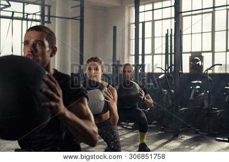 Group of three people doing squats while holding heavy medecine balls. Young men and fitness woman doing bodyweight exercises using weight ball. People in a row squatting with weighted balls at gym.