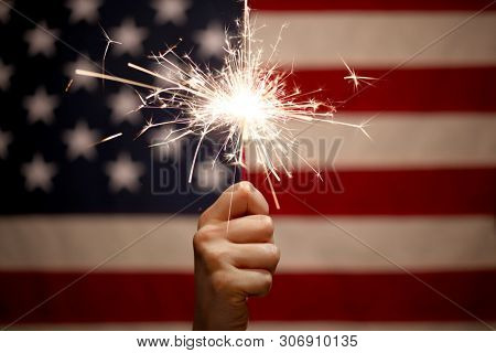 Hand holding lit sparkler in front of the American Flag for 4th of July celebration