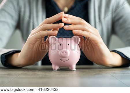 Close up of hands of woman protecting piggy bank for saving. Hands covering savings on table. Closeup of pink piggy bank protected by woman hands. Save money, investment and financial safety concept.