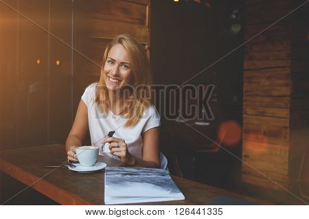 Attractive happy hipster girl with good mood posing while sitting alone in modern coffee shop interior cheerful Caucasian woman with beautiful smile enjoying her recreation time in cozy cafe bar