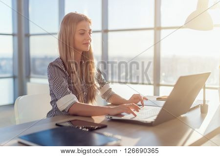 Young beautiful female copywriter typing texts and blogs in spacious light office, her workplace, using pc keyboard. Busy woman working