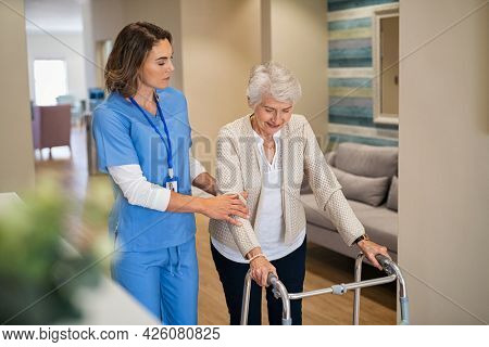 Lovely nurse helping old woman to walk at nursing home with walker. Young nurse helping senior patient using a walking frame to walk in hospital corridor. Caregiver and disabled lady in care facility.