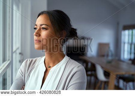 Mature african woman looking outside window with uncertainty. Thoughtful mid adult woman looking away through the window while thinking about her future business after pandemic. Doubtful lady at home.