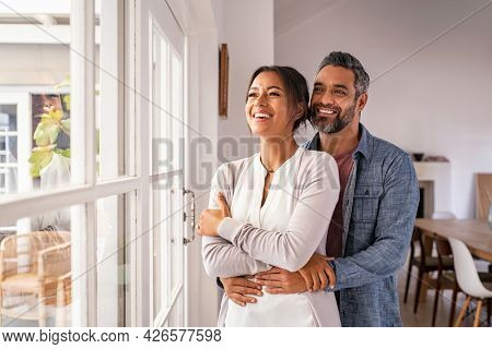 Smiling mid adult couple hugging each other and standing near window while looking outside. Happy and romantic mature man embracing hispanic wife from behind while standing at home with copy space.