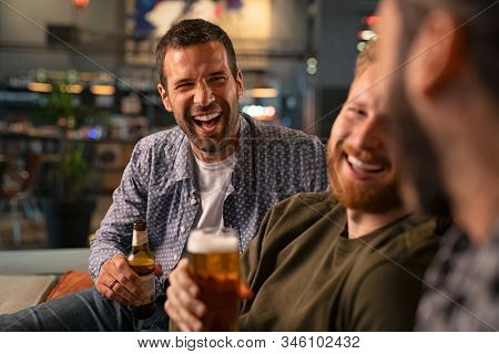 Happy young friends drinking draft beer while having fun in pub and laughing. Group of mid adult smiling men drinking beer and talk in bar while sitting on couch. guys celebrating bachelor party.