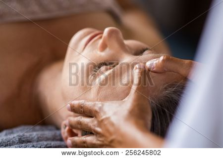 Closeup face of mature woman having facial massage at spa. Senior woman lying at spa while a massage therapist doing anti-aging treatment. Masseur doing head massage at wellness center.