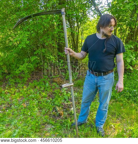 A Man Mows The Grass With A Hand Scythe. Mow The Grass With An Old Scythe. The Farmer Traditionally 