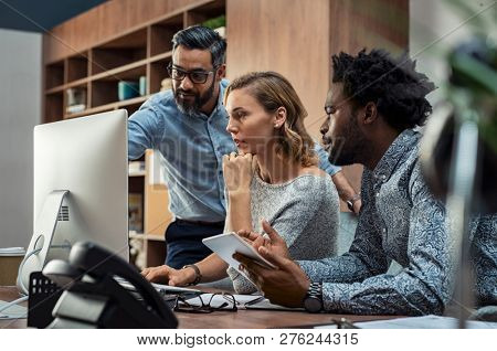 Focused businesspeople discussing project while looking at computer screen. Mature manager working with his creative team in a modern office. Multiethnic casual teamwork at work.