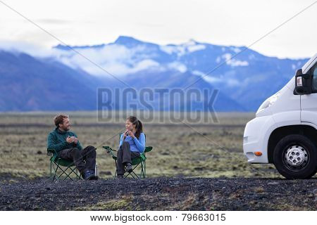 Travel couple by mobile motor home RV campervan. People sitting in chairs relaxing camping and enjoying traveling on Iceland in recreational vehicle. Young couple enjoying coffee in nature landscape.