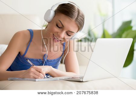 Smiling teen girl wearing headphones listening to audio course using laptop at home, making notes, young woman learning foreign languages, digital self education, studying online, enjoying music