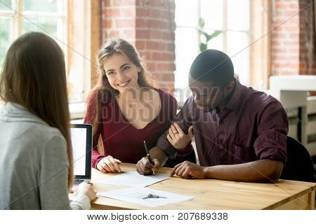 African american man signing home purchase agreement in front of realtor, his smiling wife sitting near and looking at female real estate agent. Young multiethnic family leasing new apartment concept.