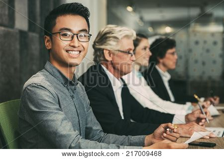 Smiling young Asian businessman wearing glasses and shirt sitting at conference table in boardroom with his colleagues, holding pen and looking at camera. Business people at workshop or seminar