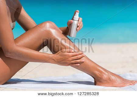 Sunscreen suntan lotion in spray bottle. Young woman in spraying tanning oil on her leg from bottle. Lady is massaging sunscreen lotion while sunbathing at beach. Female model during summer vacation.