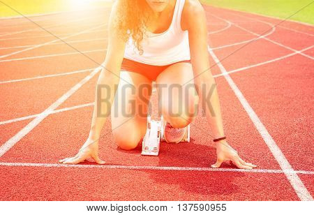 Runner woman at start line on red athletic track - Cropped girl sprinter athlete kneeling on starting block with sunrise lights background - Concept of human concentration -