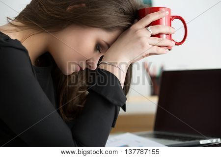 Tired female employee at workplace in office holding red cup and touching her head. Sleepy worker early in the morning. Overworking making mistake stress termination or depression concept