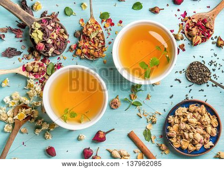 Two cups of healthy herbal tea with mint, cinnamon, dried rose and camomile flowers in spoons over blue background, top view