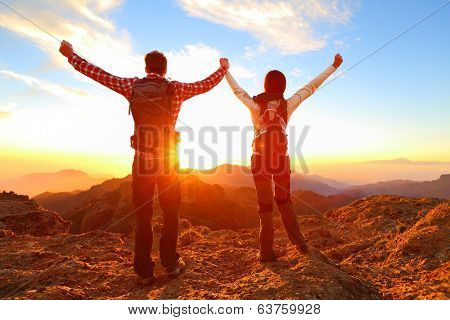 Freedom - Happy couple cheering and celebrating. Hiking man and woman raising arms excited in celebration outdoors. Hikers at sunset in mountain enjoying mountain top summit and success.