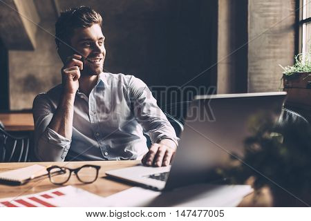 Always in touch with colleagues. Confident young man in smart casual wear talking on the mobile phone and smiling while sitting at his working place in office