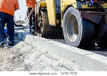 Small steamroller is flatting new road made of asphalt. Detail on newly installed curb. Close up view on roadside.